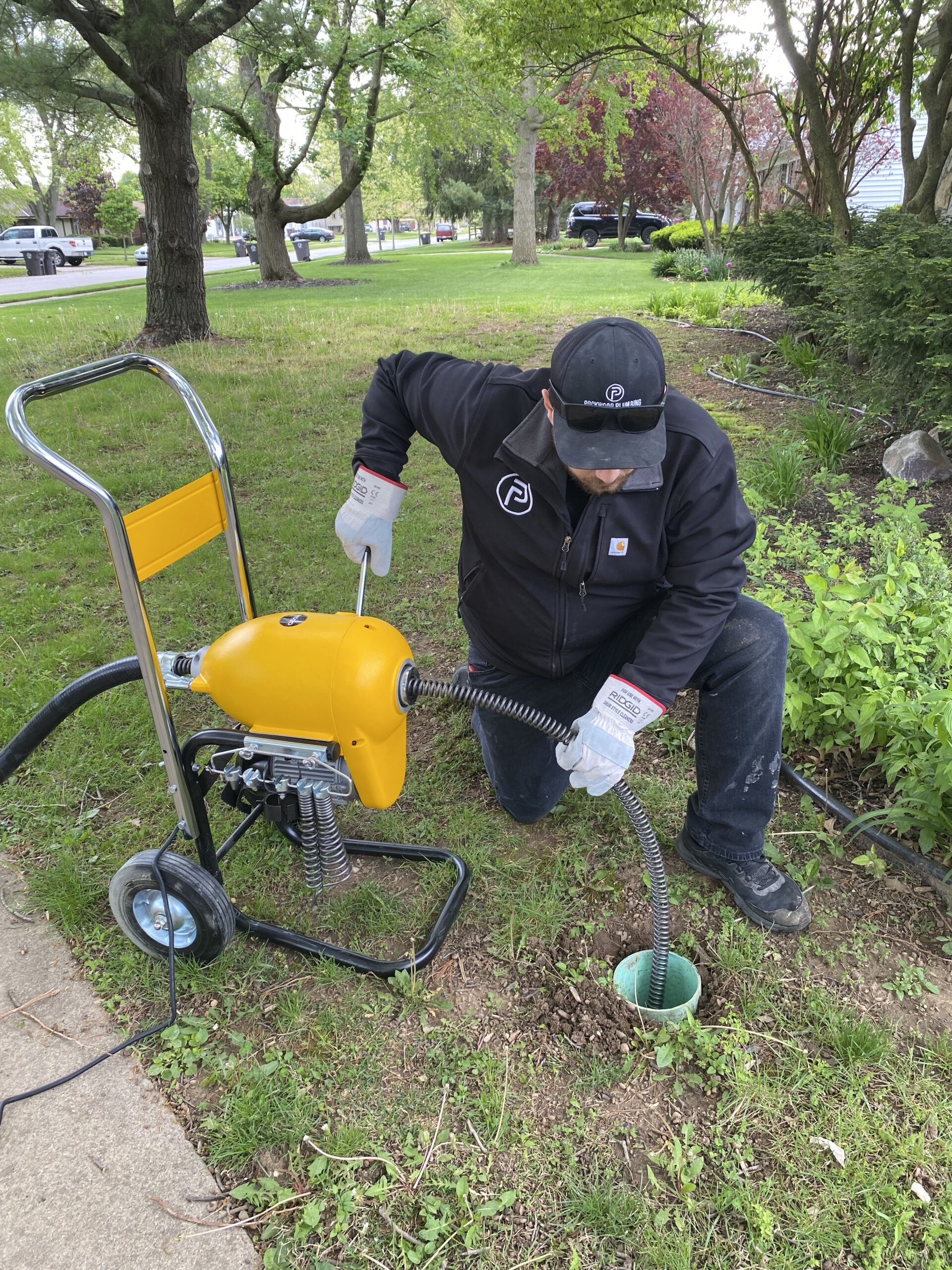 A person in work attire uses a drain cleaning machine outdoors, feeding a cable into a ground pipe surrounded by grass and shrubs.