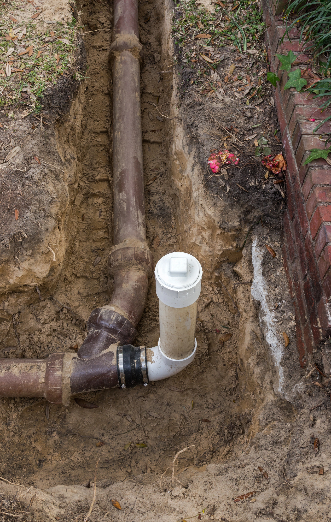 Exposed underground pipe with a vertical cleanout valve, surrounded by soil and near a brick structure, part of the sewer lines system.
