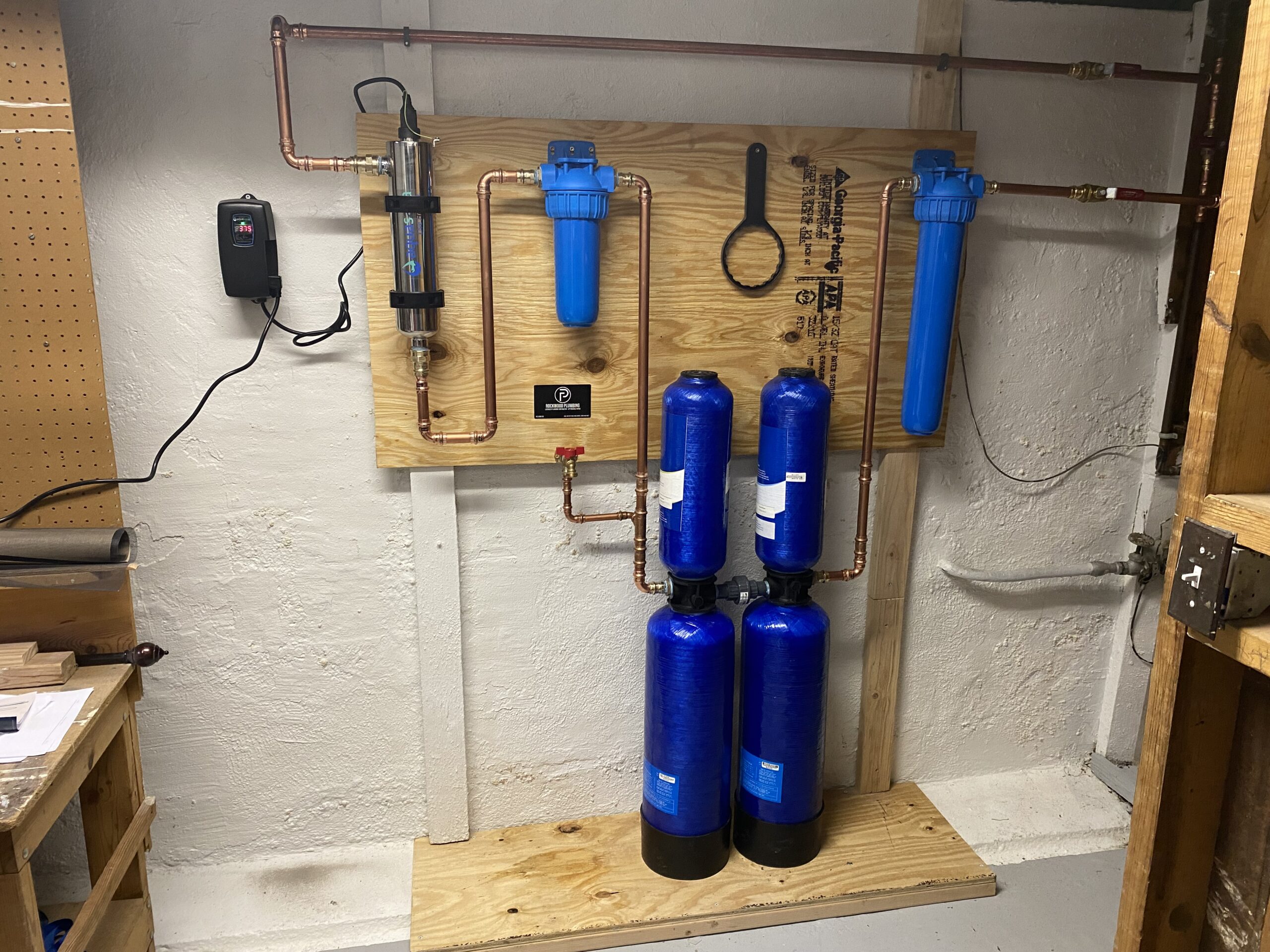 Water filtration system setup on a wooden board, featuring two blue cylindrical tanks, pipes, and filters against a basement wall.